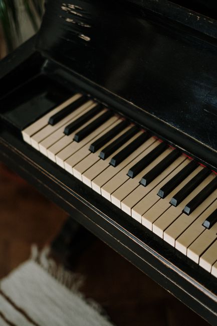 Close-up image of a black upright piano with white and black keys visible, situated indoors on a wooden floor. The piano appears to be part of a home environment, possibly in preparation for a home re