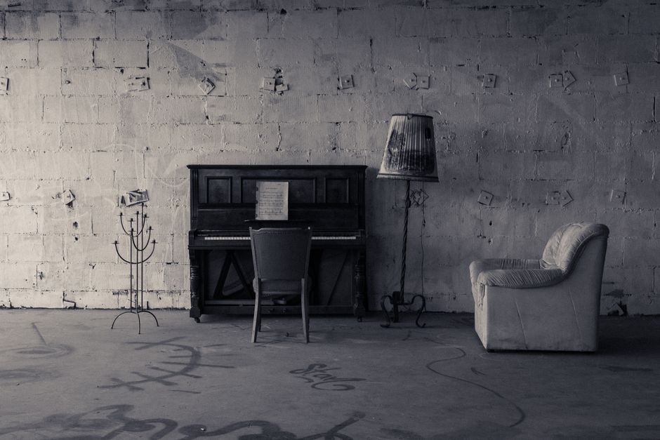 Close-up image of a black upright piano with white and black keys visible, situated indoors on a wooden floor. The piano appears to be part of a home environment, possibly in preparation for a home re